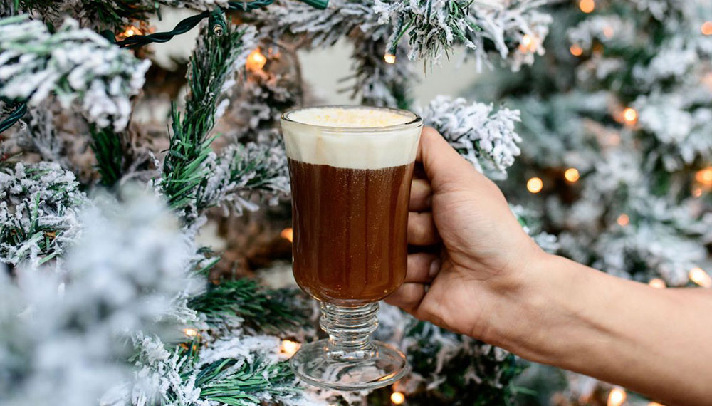 Image of hot cocoa being held in front of a Christmas tree